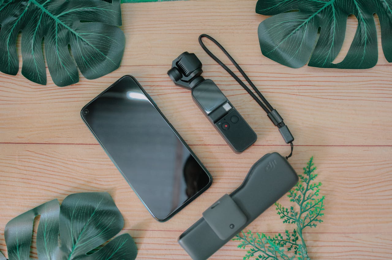 Top view composition of mobile phone placed near USB flash drive and mini gimbal camera on wooden table with green foliage